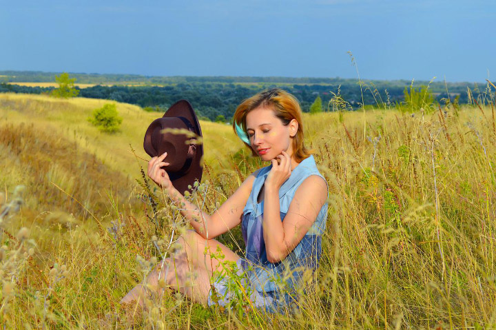 Victoria in a light blue blouse sitting amidst tall grass on a hill with a wide open sky behind her.
